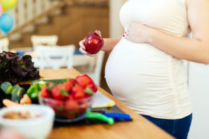 A pregnant woman holds an apple. She stands next to a counter full of fruits and vegetables.