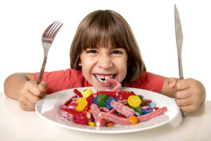 A young girl raises her eating utensils in the air and looks eager to eat some candy on her plate.