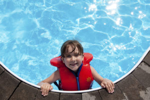 A kid poses for a photo inside a swimming pool and smiles. The kid is wearing a life vest.