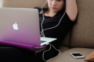 A teenage girl sits on a couch with her laptop in her lap. She wears earbuds.