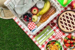 A picnic basket holds bananas, apples, and other fruit. A pie, salad, and bowl of strawberries are placed on a picnic blanket.