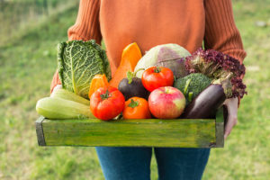 A woman appears to be holding tomatoes, eggplants, lettuce, peppers, and more vegetables in a wood bin.