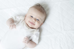 A baby boy with blue eyes lies on a white blanket and giggles.