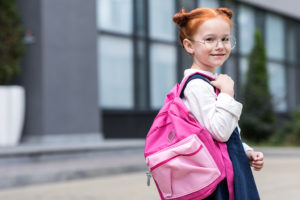 A red-haired little girl with glasses carries a pink backpack on her shoulder.