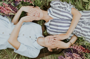 Two women lie in the grass covering each others eyes with their hands.
