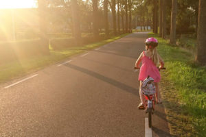 A young girl rides a bicycle along a paved path outside. She wears a pink helmet on her head.