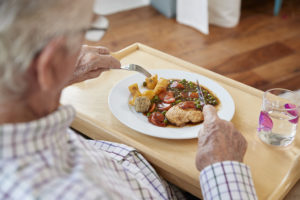 An elderly man eats a plate of chicken and vegetables with a knife and fork.