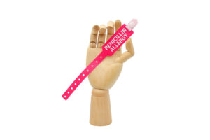 A wooden hand holds a pink snap closure wristband that says, "Penicillin Allergy."