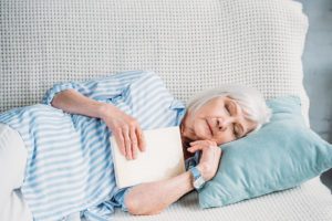 An elderly woman lies down on a couch with a book in her hand. She appears asleep.