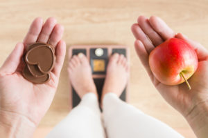 A woman stands on a scale, and looks down to see chocolate in one hand and an apple in another.