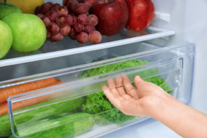 A person opens a refrigerator shelf with carrots, cucumbers and lettuce inside.