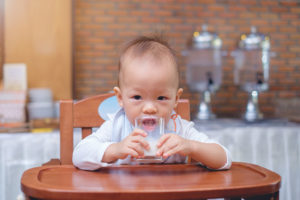 A young child sits at a feeding table and drinks a glass of milk.