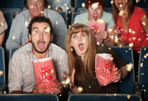 Two people holding popcorn appear to be screaming in a movie theater. They both jump and their popcorn goes everywhere.