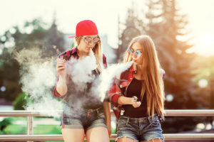 Two young ladies smoke e-cigarettes outside. 