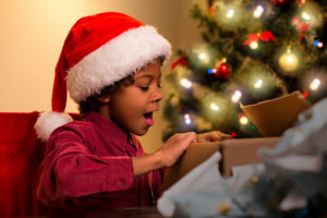 A young boy, wearing a Santa hat, opens up a present under the Christmas tree and gasps with excitement.