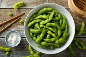 A bowl with edamame is in focus. Salt and chopsticks are next to the bowl.