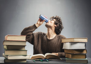 A man drinks an energy drink. He appears to be studying with multiple books.
