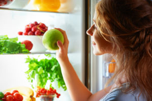 A woman looks through her fridge and grabs a green apple.