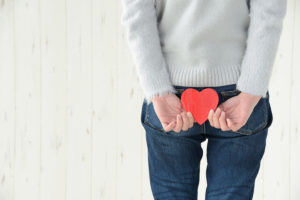 Woman wearing jeans and a gray sweater faces away from the camera and holds a red paper heart over her behind. 