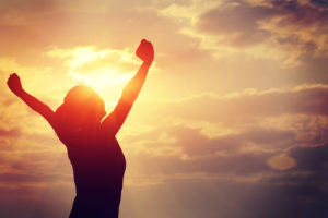 A woman raises her arms in victory as the sun rises in front of her.
