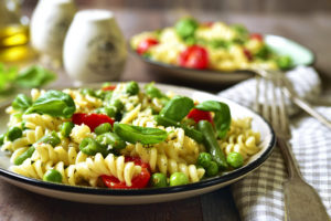 A bowl of pasta primavera is in focus. The colorful dish includes asparagus, watercress and peas.