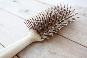 A white hair brush is shown with brown hair inside it. The brush lies on top of wood.
