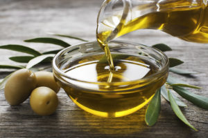 Olive oil is poured into a bowl. The bowl of olive oil is surrounded by three olives and olive leaves.