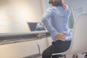 A woman sits in a chair at work and holds her back and neck with her hands. She appears uncomfortable.