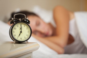 A woman sleeps in her bed. An alarm clock is shown on her nightstand.