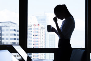 A woman stands near her work desk, holding a cup of coffee. She appears frustrated as she rests her head against her hand..