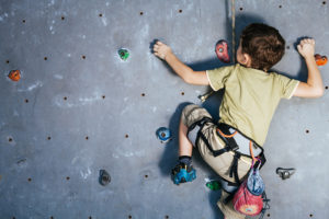 A young boy, wearing a harness, climbs up a rock climbing wall.