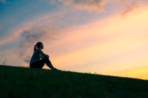 A woman sits on a hill and watches the sun set. The sky is filled with bright colors.