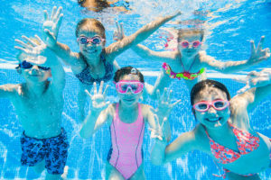 Five children pose for a photo underwater in a community swimming pool. They all wear swimsuits and goggles.
