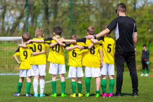 A group of young boy soccer players stand in a line and wrap their arms around each other.  They all wear yellow jerseys and colorful soccer shoes.