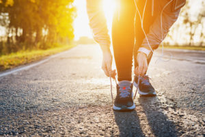 A person bends down to tie the laces on their tennis shoes before a jog outside. The sun shines down on them.