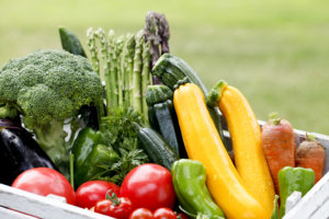 Fresh summer produce is shown in a wooden bin. Broccoli, squash, zucchini, tomatoes and carrots are shown.