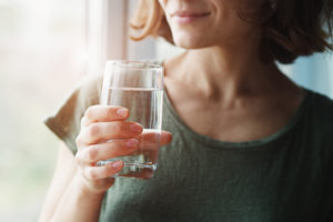 A woman holds a glass of water and looks out a window.