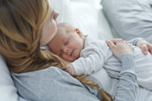 A young mom holds her newborn baby close to her chest and smiles. She lies in bed with her newborn.
