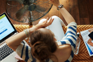 A woman sits on a couch while a fan blows air directly at her.