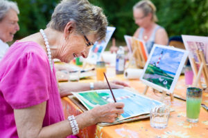 An elderly woman paints a picture of a lighthouse during a painting class.
