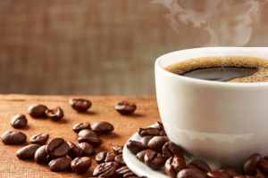 A cup of coffee sits on a table next to coffee beans.  Steam is shown above the cup of coffee.