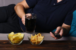 A person lounges on the couch and consumes potato chips, fries and beer.