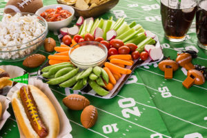 A football-themed food table is shown with a hotdog, vegetable platter, popcorn, football whistles, and mini footballs.