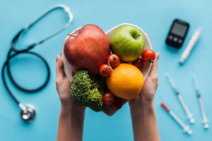 A person holds a heart-shaped bowl with apples, an orange, baby tomatoes and broccoli.  A stethoscope and insulin needles are shown in the background.