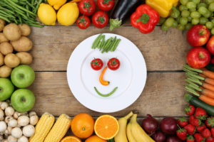 A white plate holds a few vegetables. The vegetables form a face on the plate.  The plate is surrounded by other healthy foods.