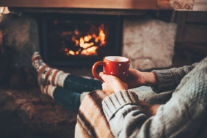A woman sits near the fireplace with a coffee cup in her hand.  She appears to be relaxing.