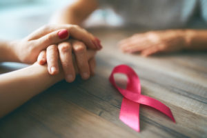 A person holds another person's hand while they sit at a table together.  A pink ribbon sits on the table next to them to symbolize breast cancer.