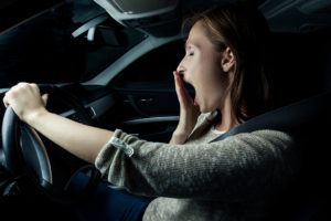 A woman covers her mouth while she yawns. She is drowsy driving.