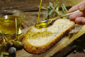 A piece of bread is shown.  A person drizzles olive oil over the piece of bread with a spoon.