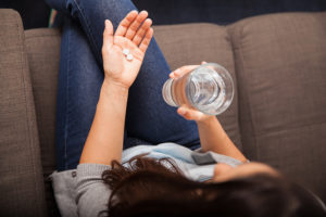 A woman sits on a couch and holds pills in one hand and a glass of water in the other hand.